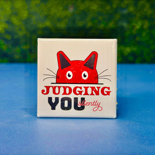 Square cat magnet featuring a red cartoon cat peeking over a ledge with the phrase “Judging You Silently” in bold red, black, and script fonts. A playful and sarcastic gift for cat lovers, introverts, or anyone with a sense of humor about being silently judged by their feline friend. Great for fridges, office boards, lockers, or as a gift for sassy pet owners and cat moms.