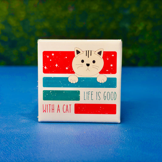 Square cat magnet featuring a sweet cartoon-style tan cat peeking over retro-styled red and teal blocks with the phrase “Life Is Good with a Cat.” A wholesome and cute cat magnet perfect for cat moms, pet lovers, and anyone who appreciates feline-themed decor. Great gift idea for birthdays, stocking stuffers, or thoughtful surprises. Ideal for fridge decor, lockers, or home offices.