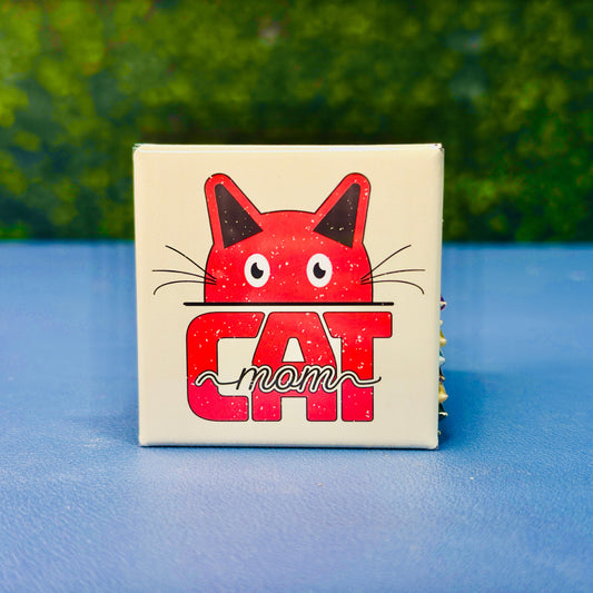 Cute square cat magnet featuring a bold red cartoon cat face peeking over the word “CAT” with the script word “mom” layered on top. Retro-inspired and lightly distressed, this magnet is the perfect gift for cat moms, pet lovers, and anyone who proudly spoils their feline friend. A fun addition to fridges, lockers, or home office boards. Great for birthdays, Mother’s Day, or just because.
