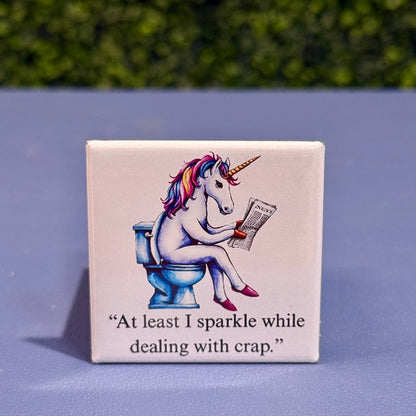 2x2 square funny unicorn magnet featuring a rainbow unicorn on a toilet reading a newspaper with the quote “At least I sparkle while dealing with crap” – adult humor fridge magnet, novelty gag gift for unicorn lovers.