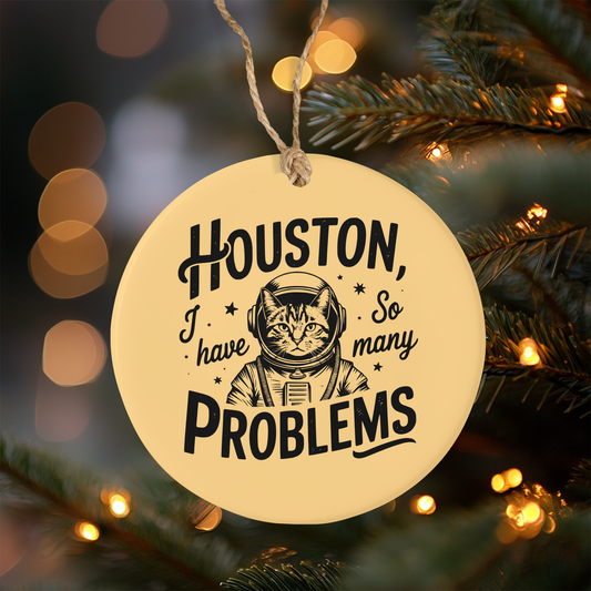 Circular ornament with a cat astronaut graphic and text 'Houston, I have so many problems' hanging on a Christmas tree.