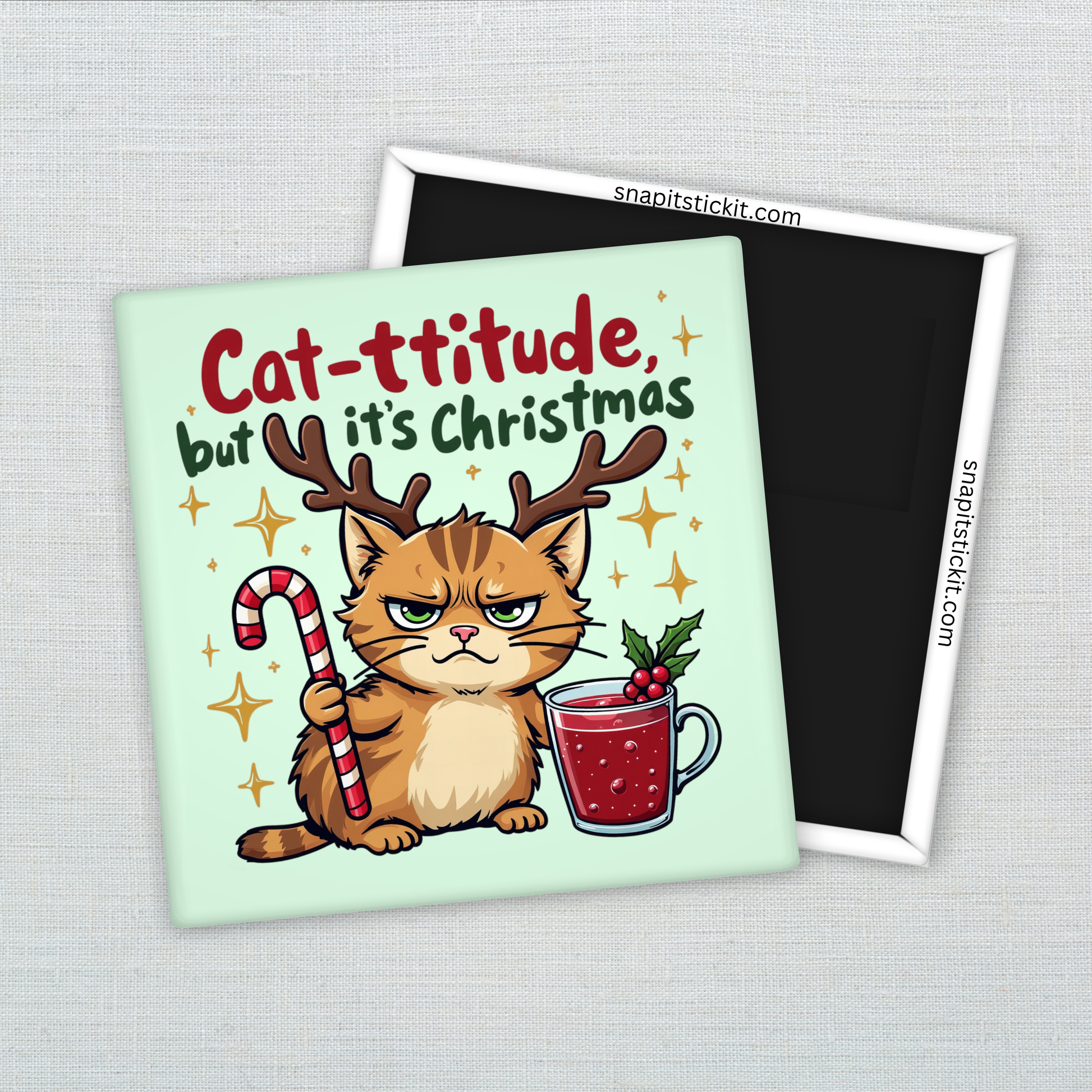 Orange cat with reindeer antlers and annoyed expression holding a candy cane, sitting beside a holiday mug. Text reads: “Cat-ttitude, but it’s Christmas.” Funny holiday 2x2 magnet.