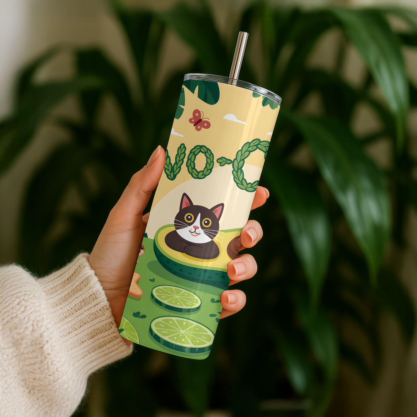 Person holding a tumbler with avocado and cat design against a blurred green plant background