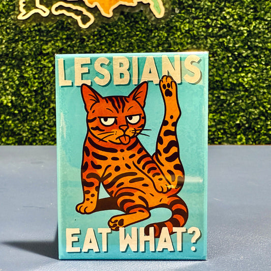 Magnet with a cat illustration and text 'LESBIANS EAT WHAT?' on a surface with a textured green background.