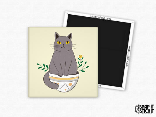 Gray cat in flower pot fridge magnet – 2x2” square magnet featuring a cartoon gray cat with yellow eyes sitting in a decorative plant pot with leaves and a small flower. Cream background with strong magnetic backing and Snap It Stick It branding. Great for cat lovers, plant moms, and whimsical decor fans.