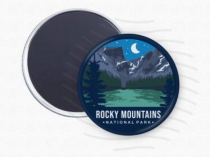 Round magnet of Rocky Mountains National Park featuring snow-capped peaks under a crescent moon, with a green alpine lake and dark pine trees in the foreground